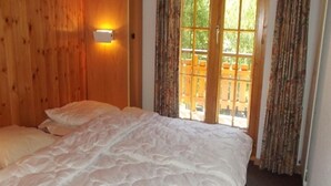 A bedroom with a wooden headboard, a large bed with white bedding, a wall-mounted light, and a view of an outdoor deck through a window. - Gorgeous Apartment Amidst Valleys (Sion)