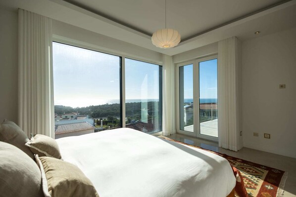 A bedroom with a large bed, a view of a cityscape through floor-to-ceiling windows, and a hanging light fixture.