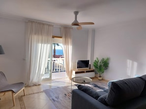 A living room with a sofa, a TV, a ceiling fan, and a view to the outside.