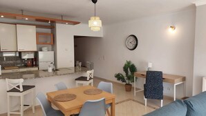 Une cuisine moderne avec des meubles blancs, un coin repas avec une table et des chaises en bois, un salon avec un canapé bleu et une horloge murale.