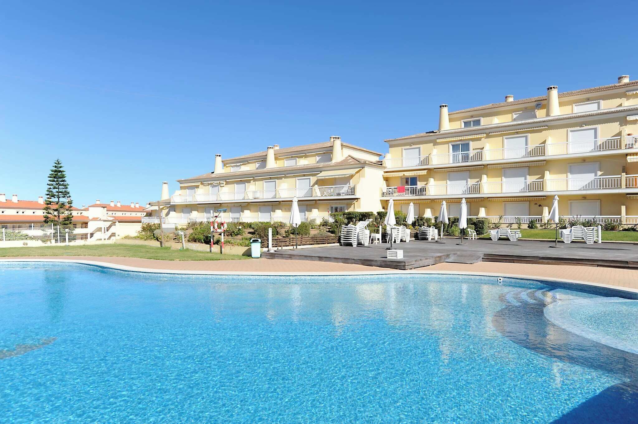 Une piscine à l’eau d’un bleu limpide, entourée d’une zone dallée avec des chaises longues et des parasols. Au fond, on aperçoit des immeubles à plusieurs étages dotés de balcons.
