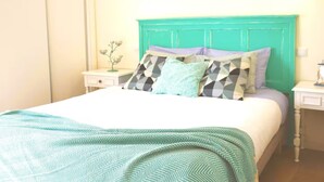 Een bed met een beddekleed met een patroon, een blauwgroen hoofdeinde en meerdere kussens met verschillende ontwerpen.