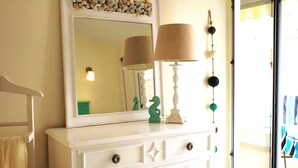 Een witte ladekast met er boven een spiegel, een lamp, decoratieve objecten, en uitzicht op het buiten.