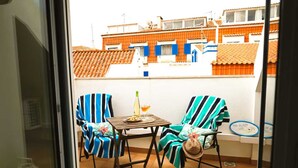Een balkon met twee gestreepte stoelen, een klein tafeltje met een fles en glas, en een uitzicht op omliggende gebouwen.