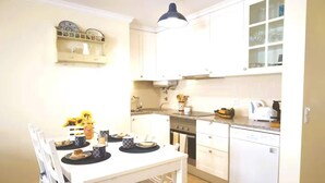 Een kleine keuken met witte kastjes, een eettafel gedekt met borden en kopjes, en een koffiezetapparaat.