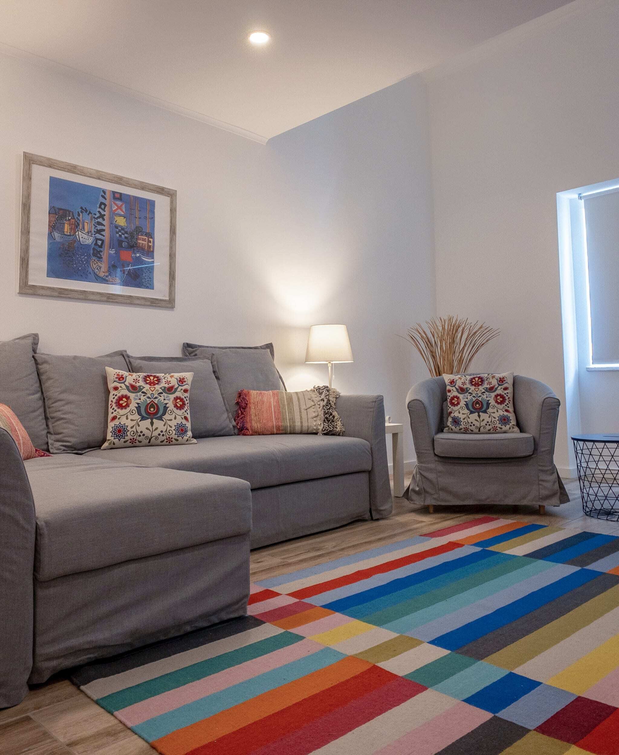 Ein Wohnzimmer mit einer grauen Sofagarnitur, einem farbenfrohen StreifenTeppich, einer Lampe und einem gerahmten Bild an der Wand.
