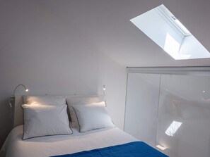 A modern bedroom with a skylight, a bed with white bedding, and a blue accent.