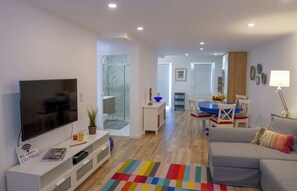 A modern living room with a flat-screen TV, a white entertainment unit, a colorful rug, a gray sofa, and a dining area with a blue table and chairs.