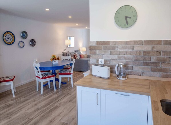 A modern kitchen with a brick wall, a dining area with a blue table and white chairs, and a wall clock.