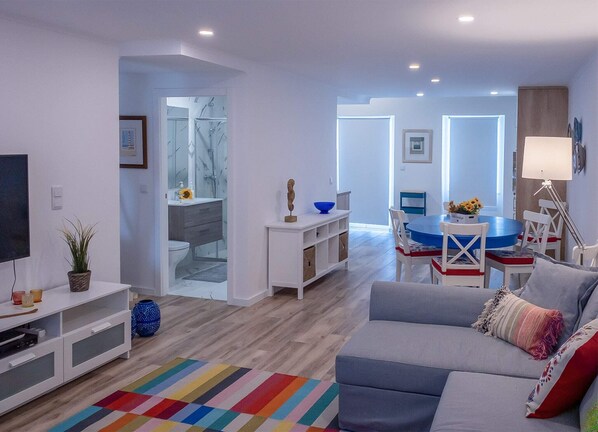 A modern living room with a grey sofa, a colorful striped rug, a dining area with a blue table, and a white TV stand.