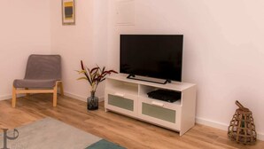 A living room with a flat-screen TV on a white cabinet, a grey sofa, a small table with a vase of flowers, and a wooden floor.