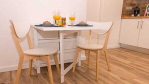 A small dining area with a white table and two chairs, a coffee machine, and a fruit bowl on the counter.