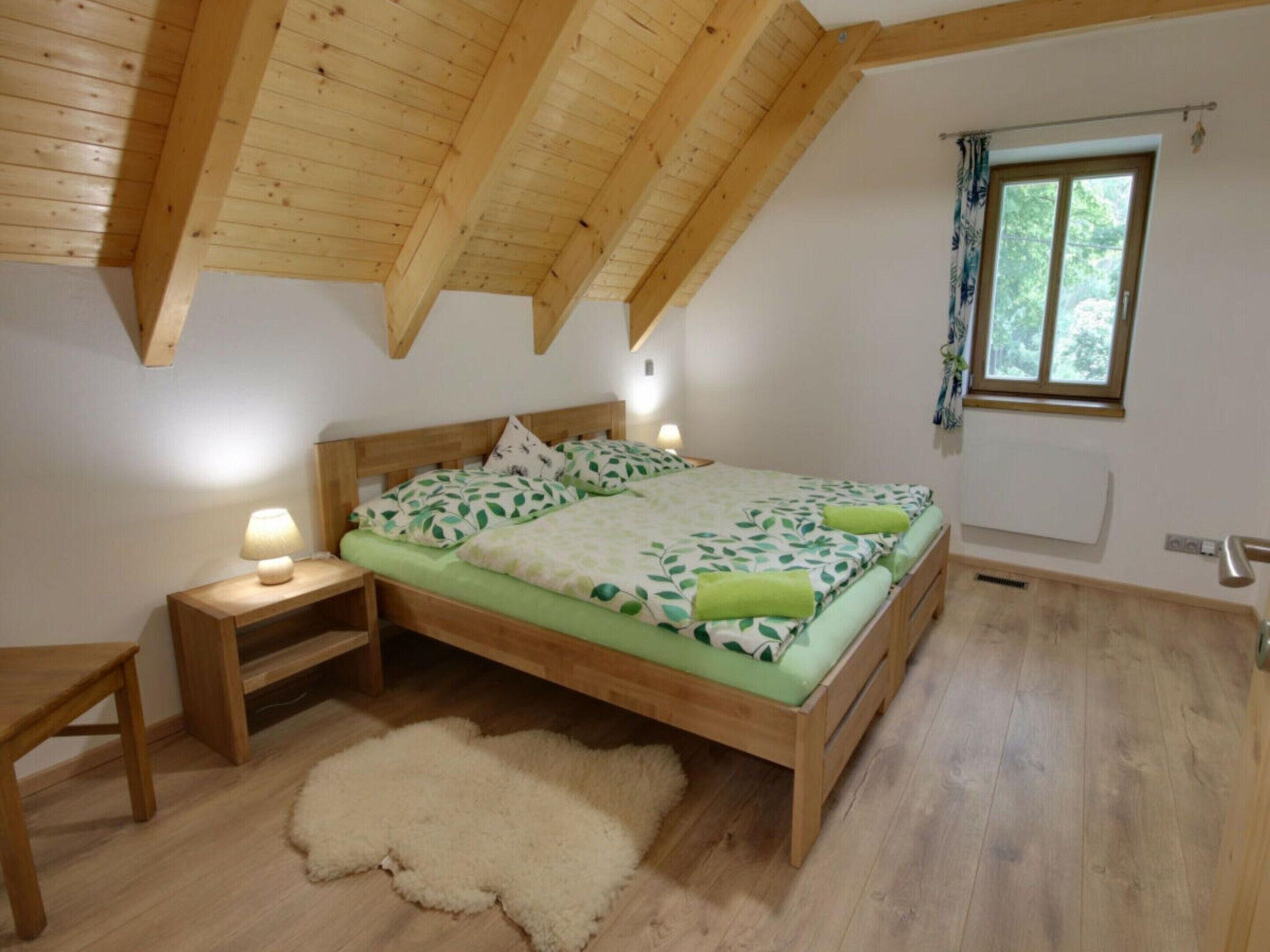 A bedroom with a wooden ceiling, a bed with green bedding, a bedside table, a nightstand, a fluffy rug, and a window with curtains.