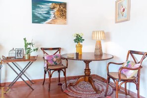 A dining area with a round wooden table, two chairs, a lamp, and a painting of a beach. - Window to the Valley (Mafra)