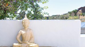 A seated Buddha statue against a white wall with a backdrop of trees and a distant building. - Window to the Valley (Mafra)