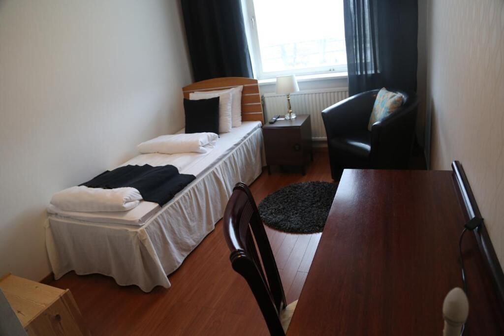 A small, neatly arranged bedroom with a single bed, a chair, a table, and a window with curtains.