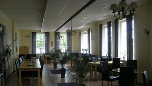 Ένας ευρύχωρος εσωτερικός χώρος με πολλά τραπέζια και καρέκλες, γλάστρες με φυτά  και μεγάλα παράθυρα που επιτρέπουν την είσοδο φυσικού φωτός.