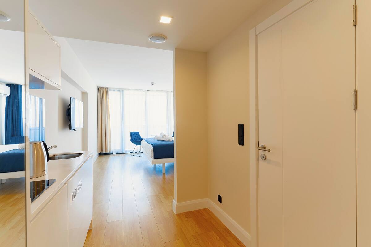 Una habitación de hotel moderna con kitchenette, una cama con cabecera azul, una zona de estar con un sillón azul y un ventanal con cortinas.