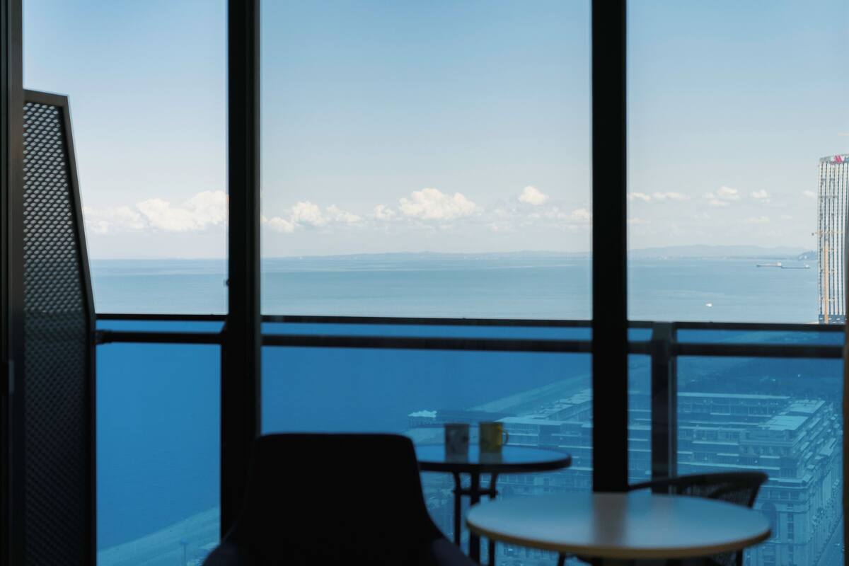 Habitación con amplia ventana que da a un paisaje urbano y una masa de agua. Hay dos sillas y una mesita.