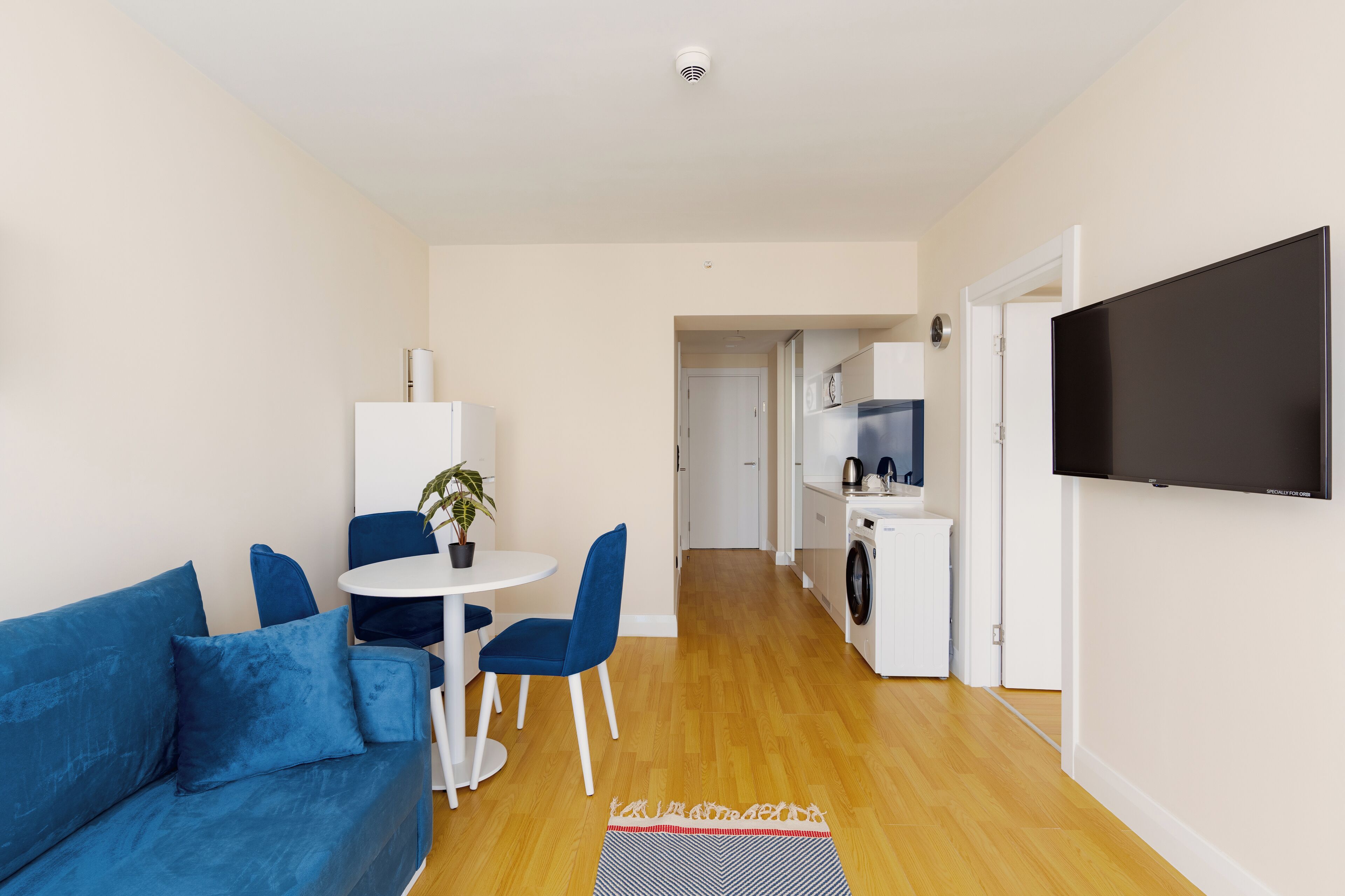A modern living room with a blue sofa, a dining table with chairs, a small potted plant, and a TV mounted on the wall.