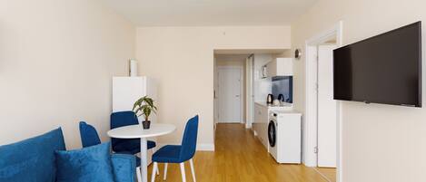 A modern living room with a blue sofa, a dining table with chairs, a small potted plant, and a TV mounted on the wall.