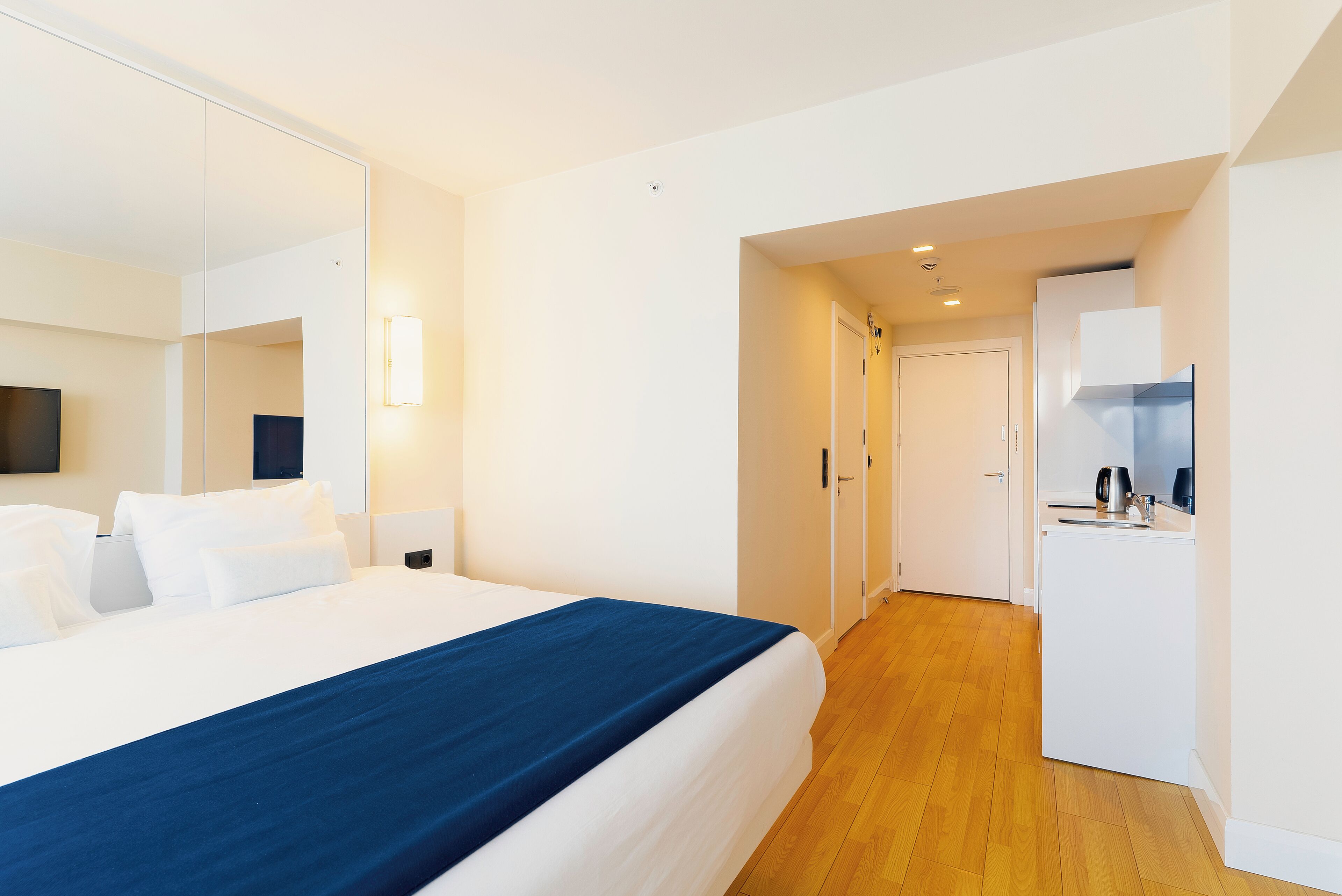 A modern hotel room with a large bed, a small kitchenette, and a mirror.