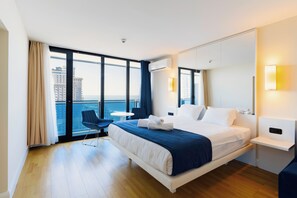 A modern hotel room with a large bed, a small desk, a chair, and a balcony with a city view.