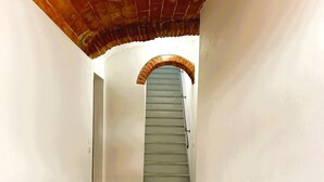 A narrow hallway with a tiled floor, a wooden door on the left, and a staircase with a brick archway leading to a landing.
