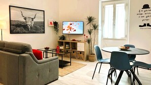 A modern living room with a grey sofa, a TV, a dining table with chairs, and a window with a view.