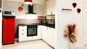 A modern kitchen with white cabinets, a red refrigerator, a black stove, and a microwave. There is a red pendant light, a clock, and decorative hearts on the wall.