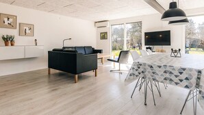 Una sala de estar moderna con un sofá negro, una mesa de comedor con sillas, un televisor y un ventanal con vistas al exterior.