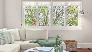 Una sala de estar con un sofá color beige, una mesa redonda de centro con libros y un control remoto, una manta verde de punto, un jarrón con una planta, y una ventana que ofrece vista a árboles.