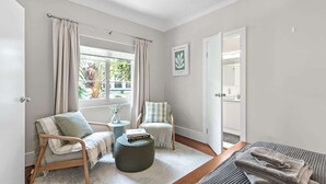 Una habitación con cama, una silla, una mesita, una ventana con cortinas y una puerta que da a otra habitación.