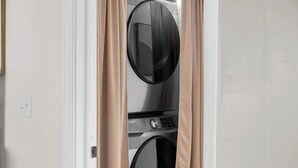 Vista frontal de uma lavanderia com máquina de lavar e secadora de abertura frontal, cortinas bege e uma tomada visível na parede.