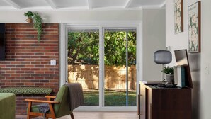 Um quarto com parede de tijolos, porta de vidro deslizante para um quintal, um sofá verde, uma cadeira de madeira e um armário de madeira escura com uma luminária.
