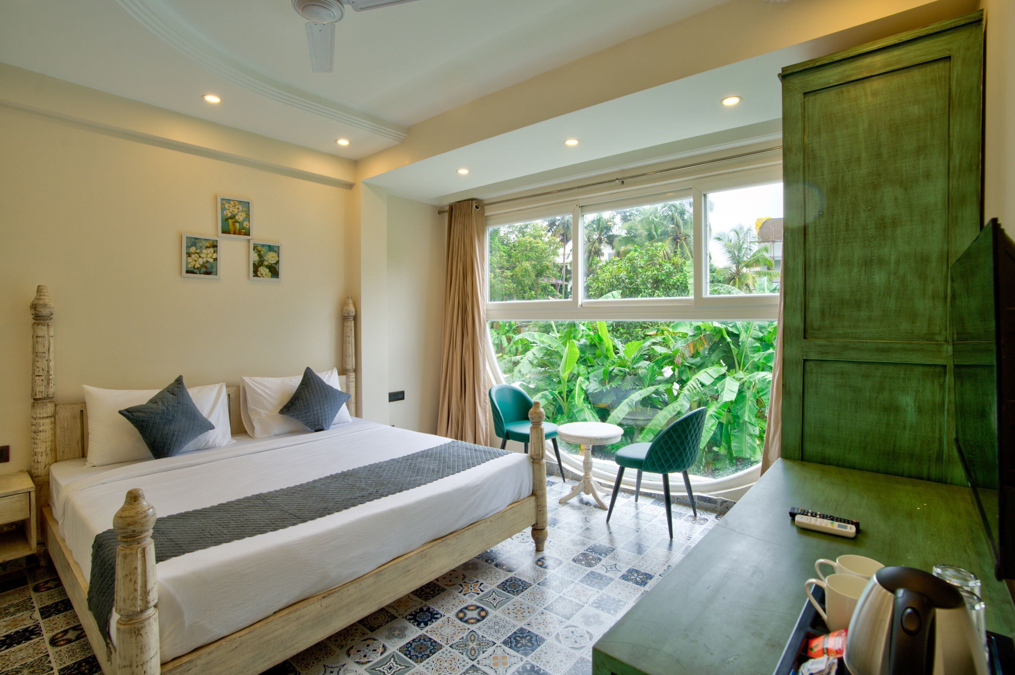 A hotel room with a bed, a desk, a chair, a window with a view of greenery, and a green cabinet.
