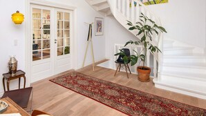 Uma sala de estar ampla, com uma grande janela, escada, tapete vermelho estampado e um vaso com planta.