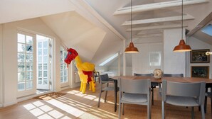 Um quarto espaçoso com teto alto, grandes janelas, área de refeições com mesa e cadeiras de madeira, e uma escultura colorida de um cavalo.