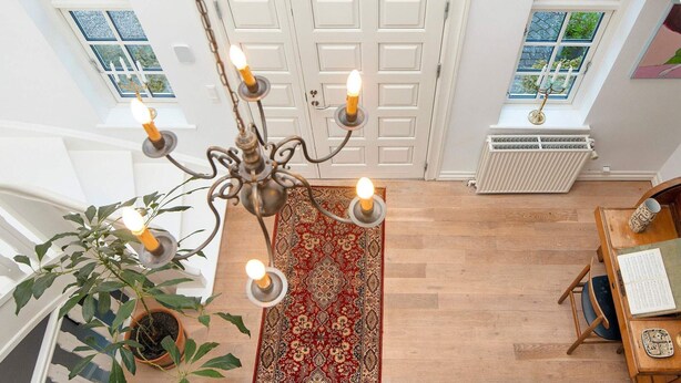 Um quarto espaçoso com um candelabro, uma grande planta em vaso, um tapete vermelho estampado e uma escrivaninha de madeira com um livro e um objeto decorativo.