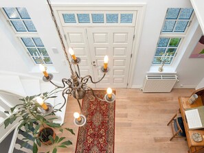 Una habitación amplia con un candelabro, una planta grande en maceta, un tapete rojo estampado, y un escritorio de madera con un libro y un objeto decorativo.