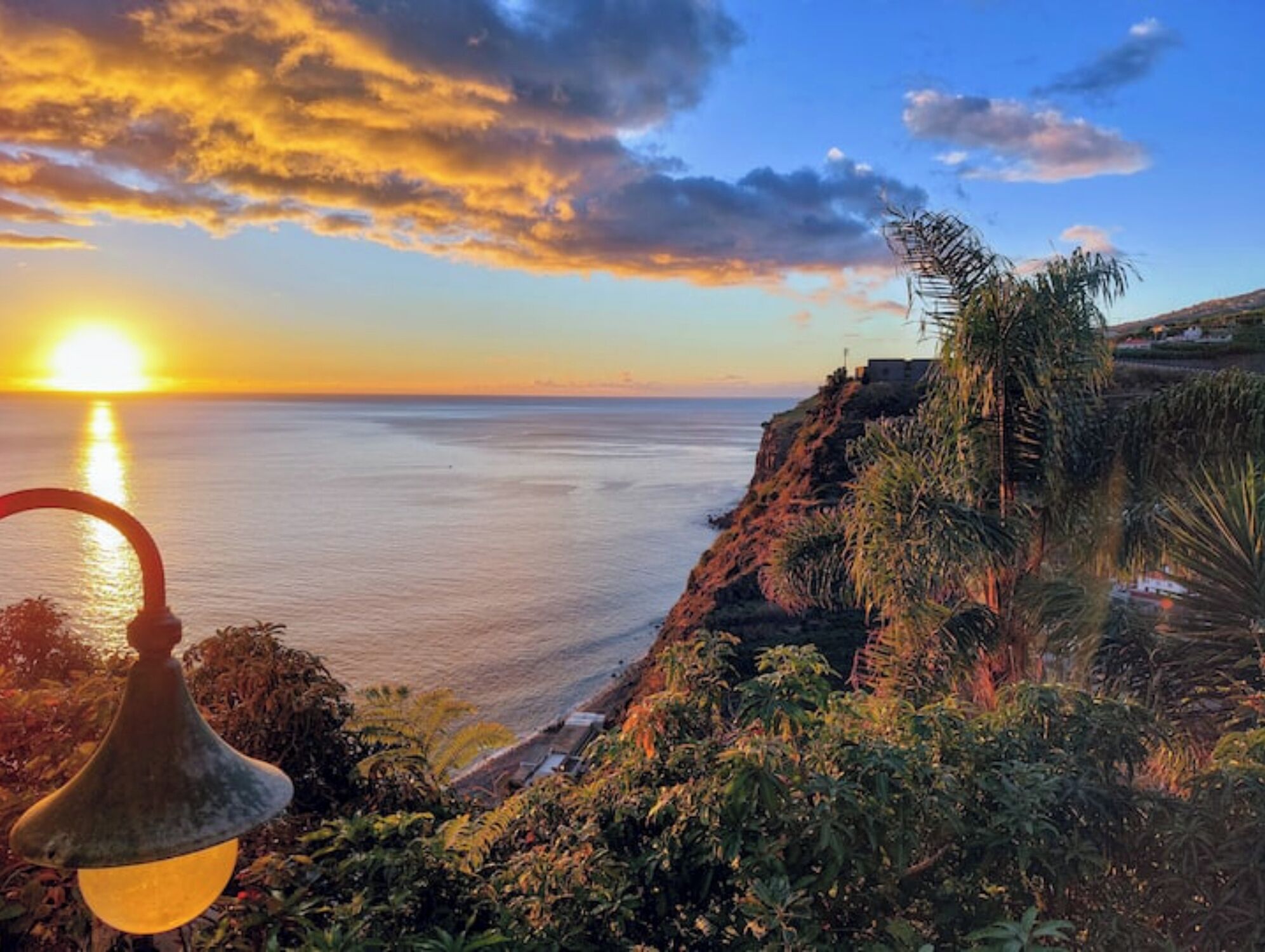 Une vue sur le coucher de soleil depuis un balcon donnant sur une zone côtière, avec de la végétation et un lampadaire.