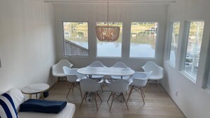 A room with a wooden floor, white walls, a white sofa with blue stripes, a round table with white chairs, and large windows overlooking a body of water. - Peaceful Lake Retreat: Modern Luxury, Comfort, Scenic Views & Paddle Boards (Austin)