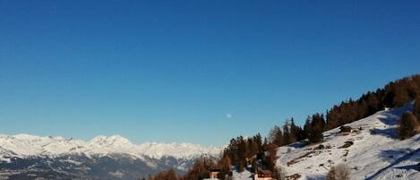 A snow-covered mountain landscape with a clear sky, a small cabin, and a distant mountain range.
