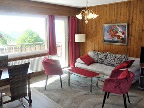 A cozy living room with a sofa, two chairs, a glass coffee table, a chandelier, a painting, and a view through a window.