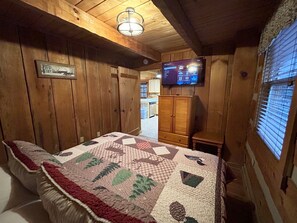A cozy cabin room with a wooden ceiling, a quilted bed, a television mounted on the wall, and a window with blinds.