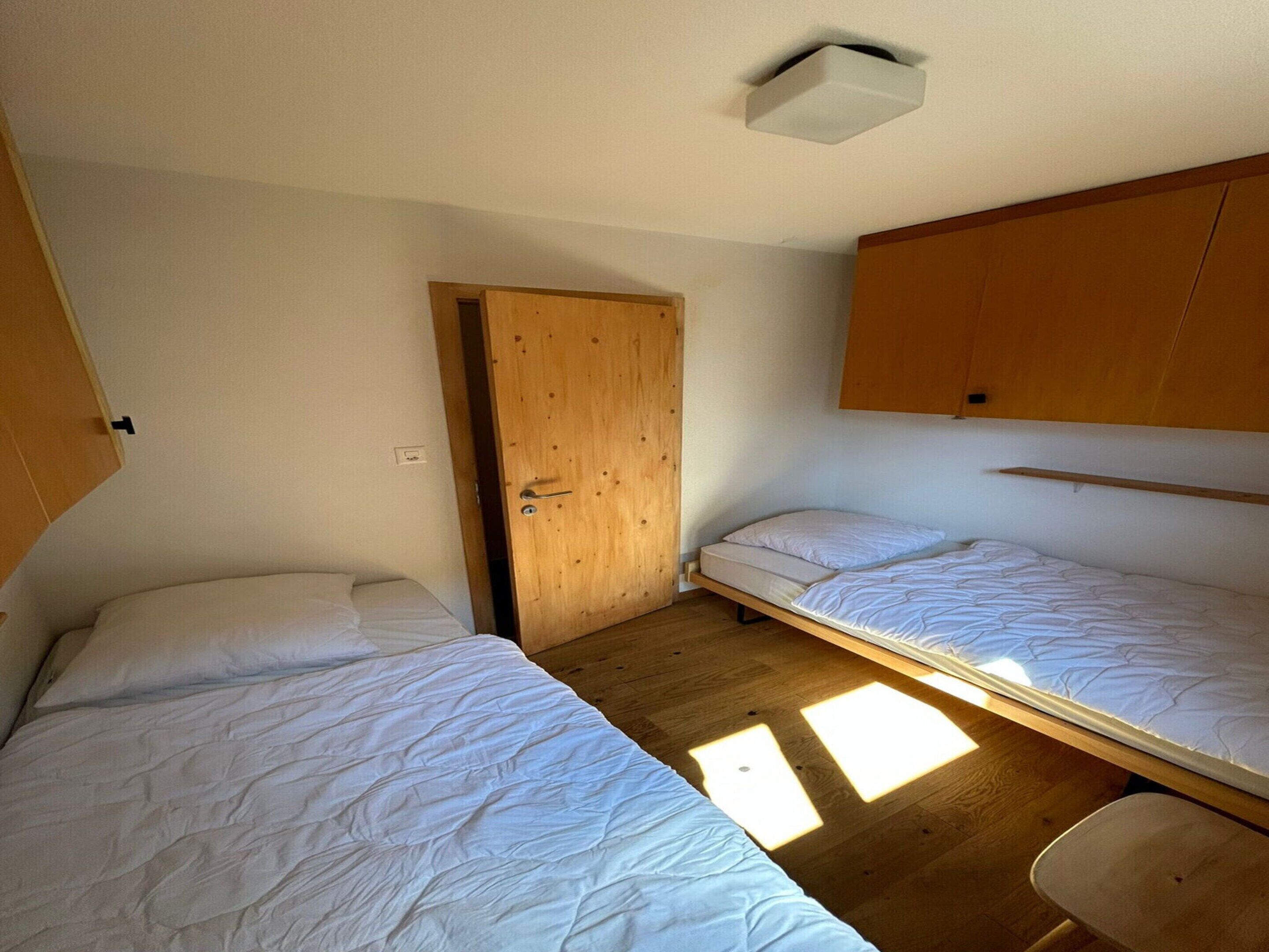 Une petite chambre avec deux lits simples, une porte en bois et une grille d’aération au plafond.