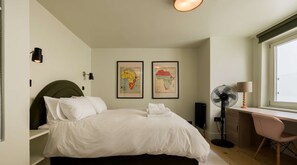 Ein ordentlich eingerichtetes Schlafzimmer mit einem Bett, einem Schreibtisch, einem Stuhl, einem Standventilator und zwei gerahmten Karten an der Wand.