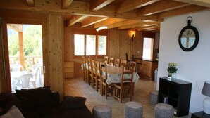 A wooden cabin interior with a dining area, a living room with a sofa and ottomans, and a kitchen area with a clock on the wall. - Chalet Nestled in Mountains and Garden (Nendaz)