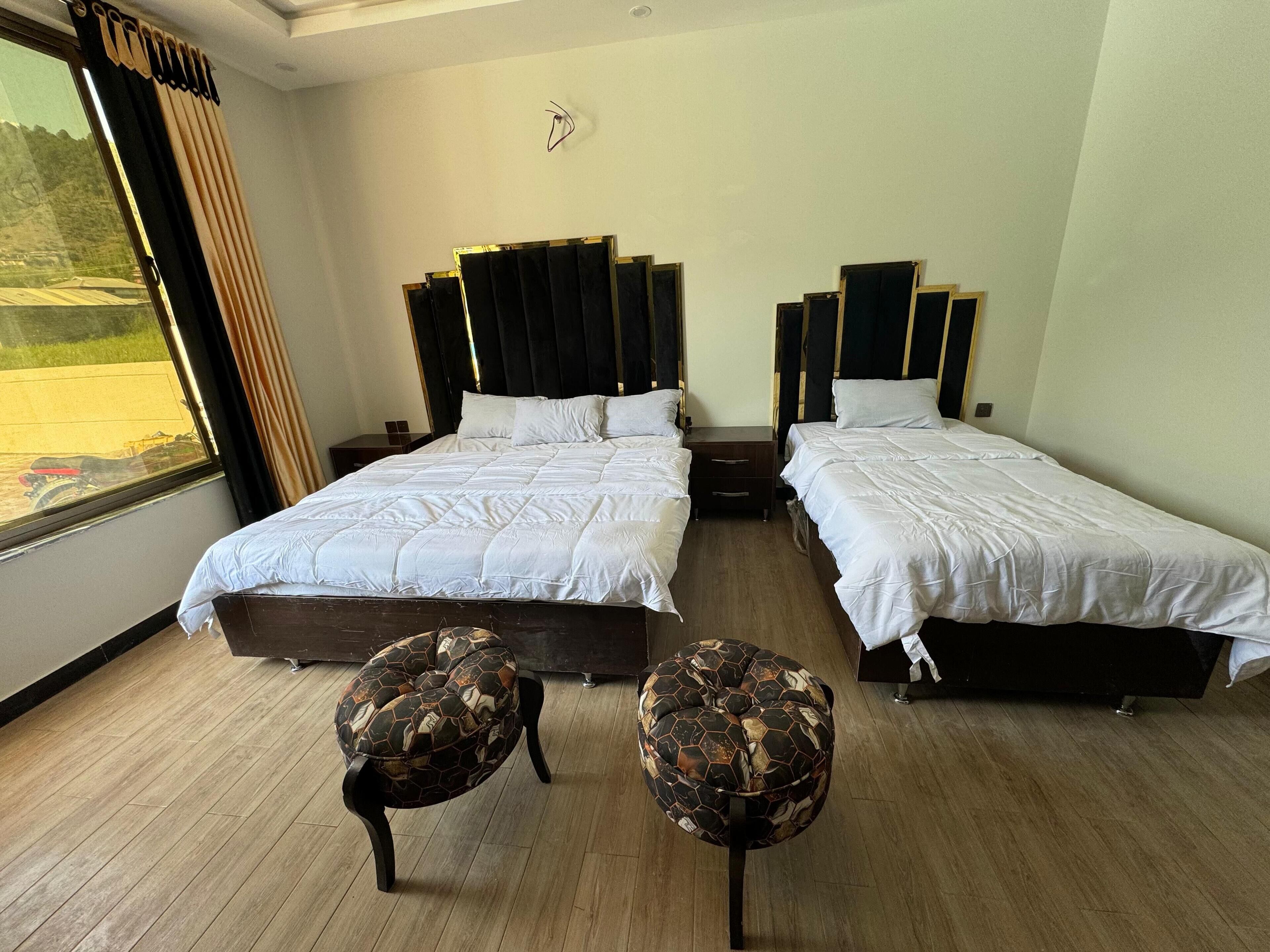 A hotel room with two beds, a window with curtains, and two ottomans.