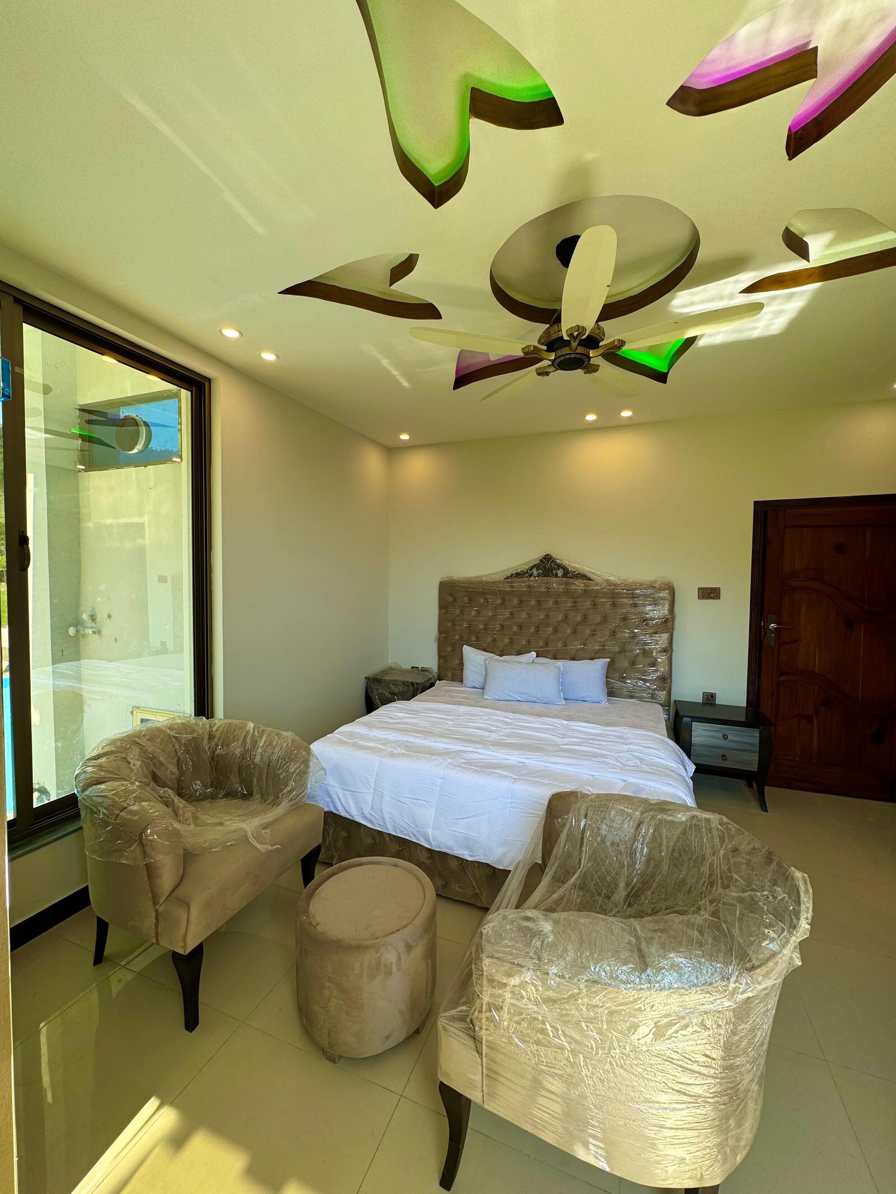 A modern bedroom with a large bed, two armchairs, a round ottoman, a ceiling fan, and a large window.